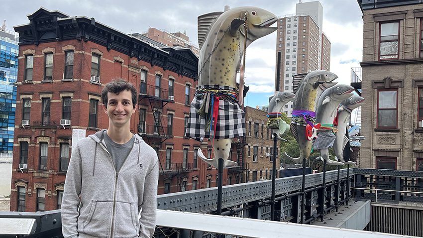 Ben stands on a roof top deck in a city with buildigs behind him