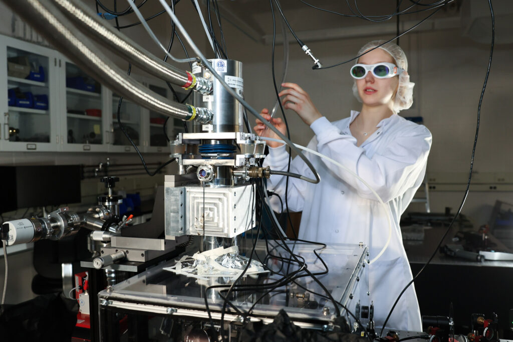 An undergraduate student wearing safety goggles, lab coat, and hair net works in the Disa photonics lab