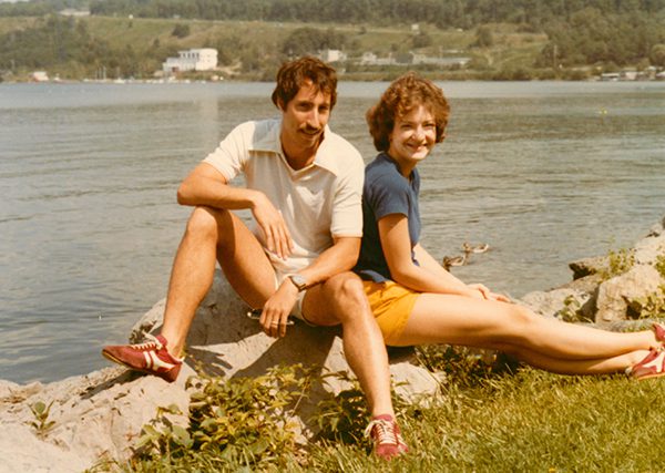 In a photo from the 1970s, a college-aged man (David Fischell) and his girlfriend sit on rocks by a lake.