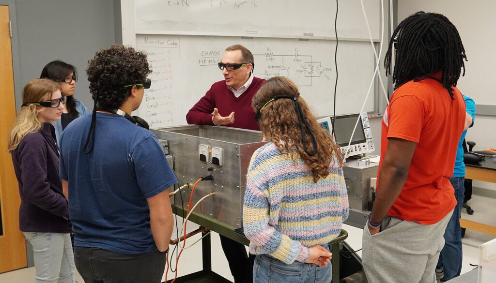 students wearing goggles are gathered around a piece of equipment where a man wearing goggles is describing it to them.