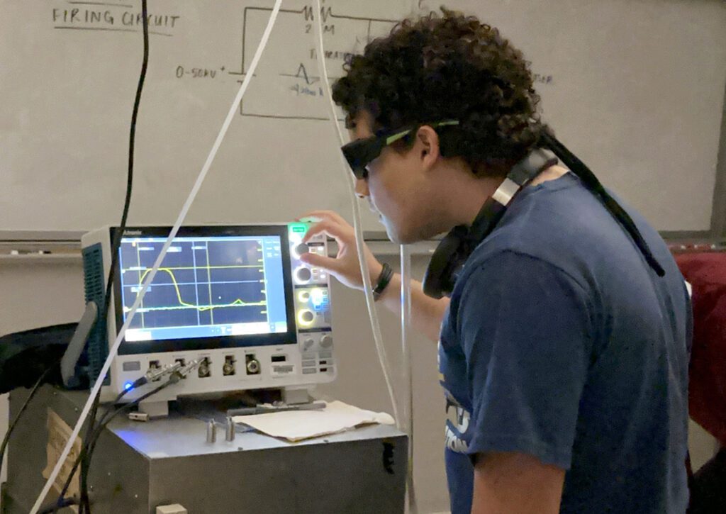 a student wearing goggles adjusts the dials on an oscilloscope that displays a wave form.