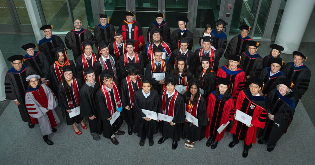 A group of of about 50 students and professors in academic robes look up at the camera.