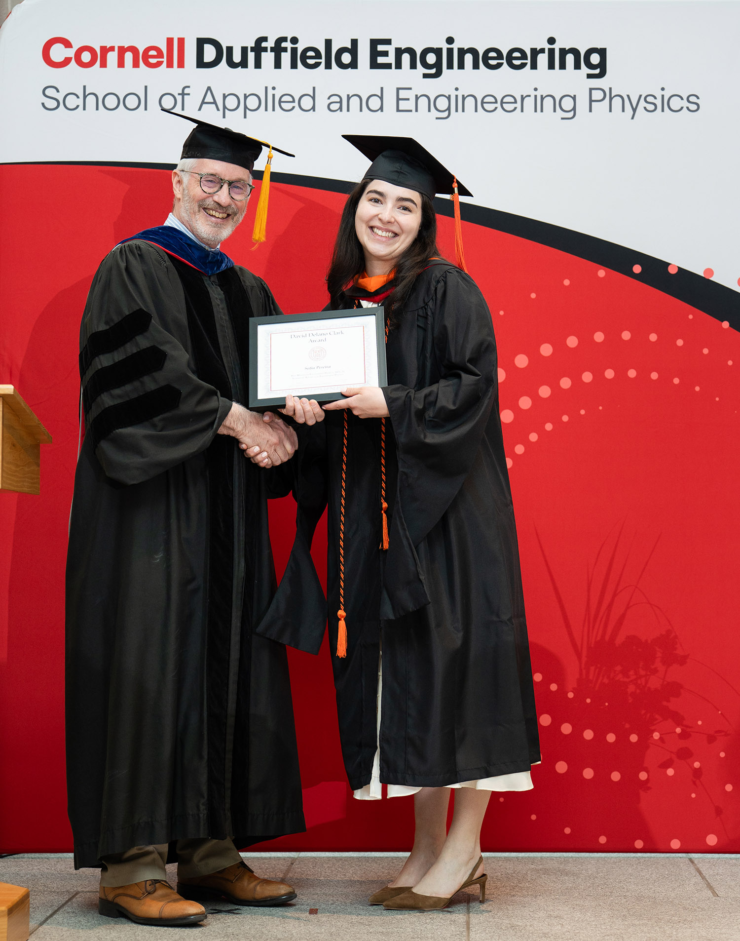 Professor Joel Brock hands an award to Sofia Pereira, who wears graduation robes.
