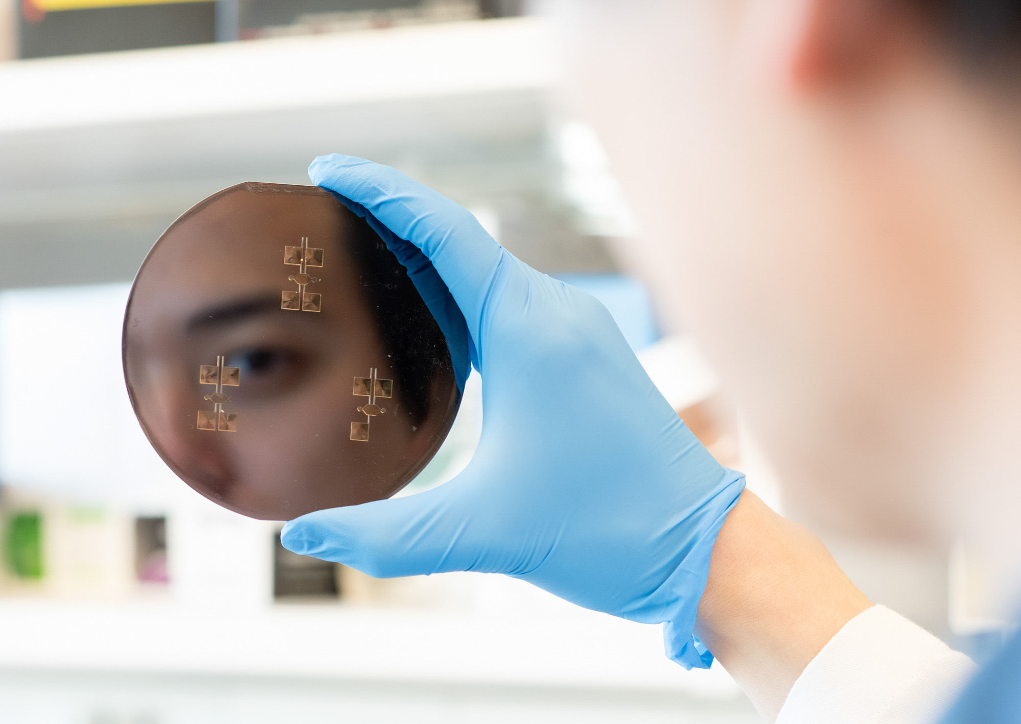 Student face reflected in silica wafer device in gloved hand in Weill Hall lab.