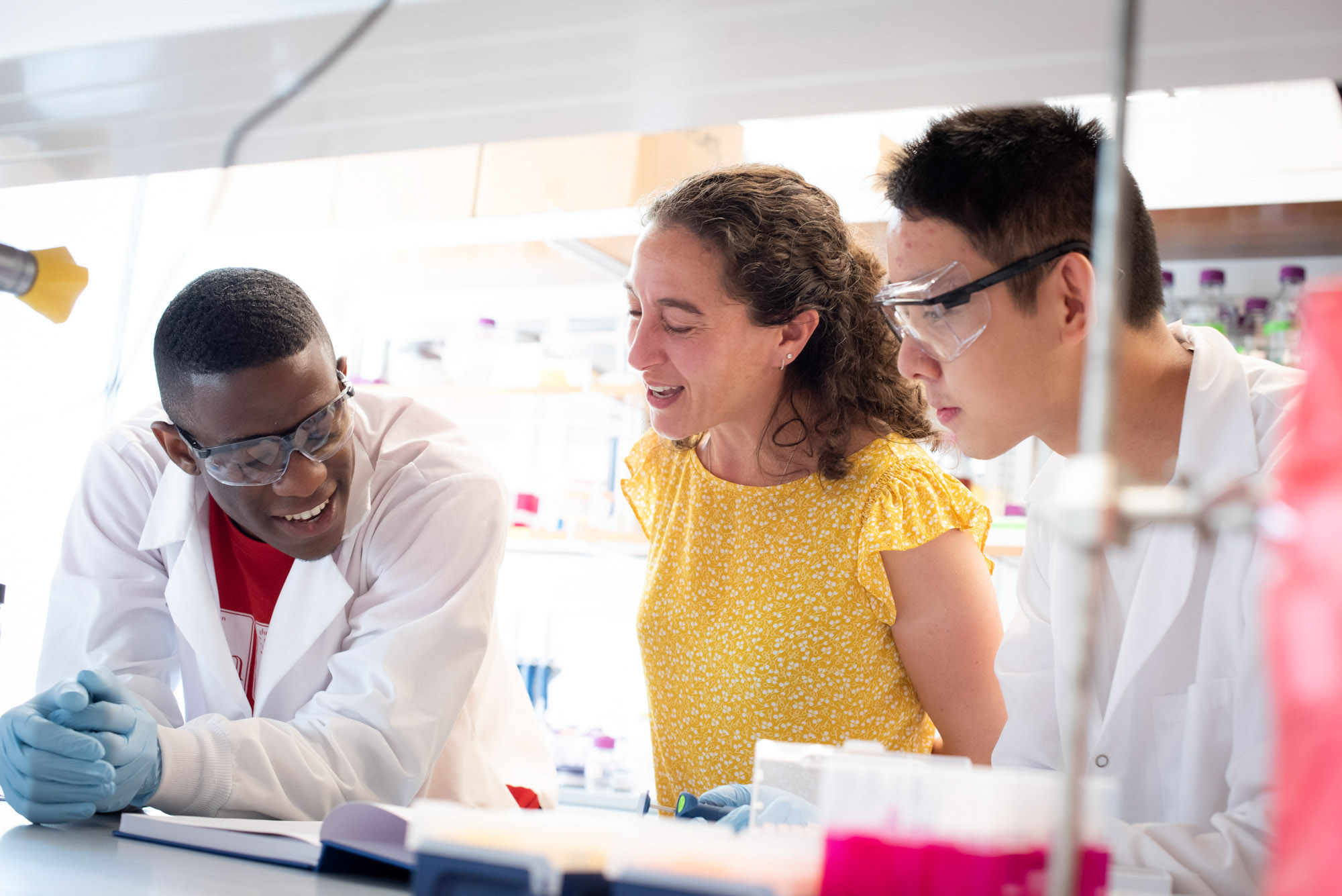 Two researchers wearing a white lab coat and safety goggles look at a book with professor Ilana Brito in the lab