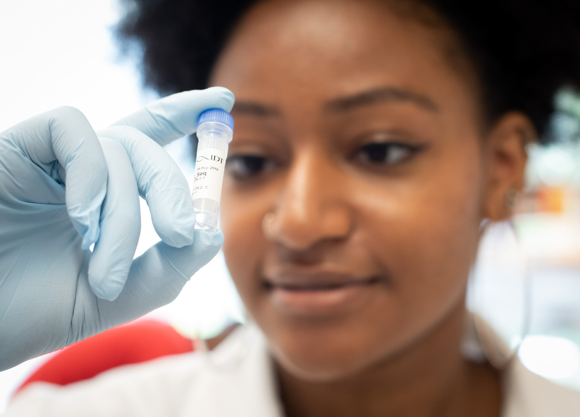 A researcher wearing a white lab coat and blue gloves examines a vial in the Brito Lab