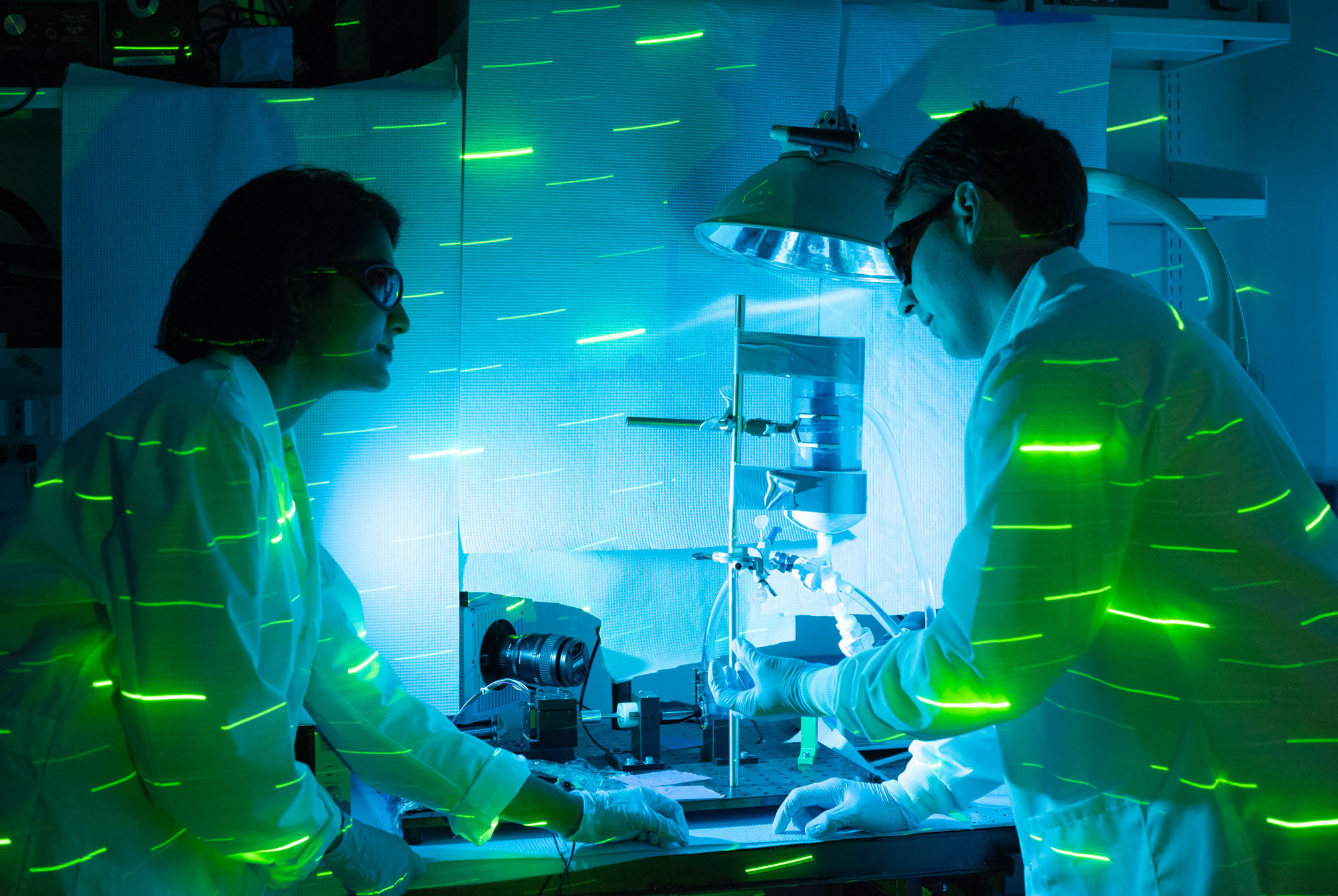 Two scientists in lab coats and safety glasses work in the Antaki lab with green laser beams illuminating the scene.