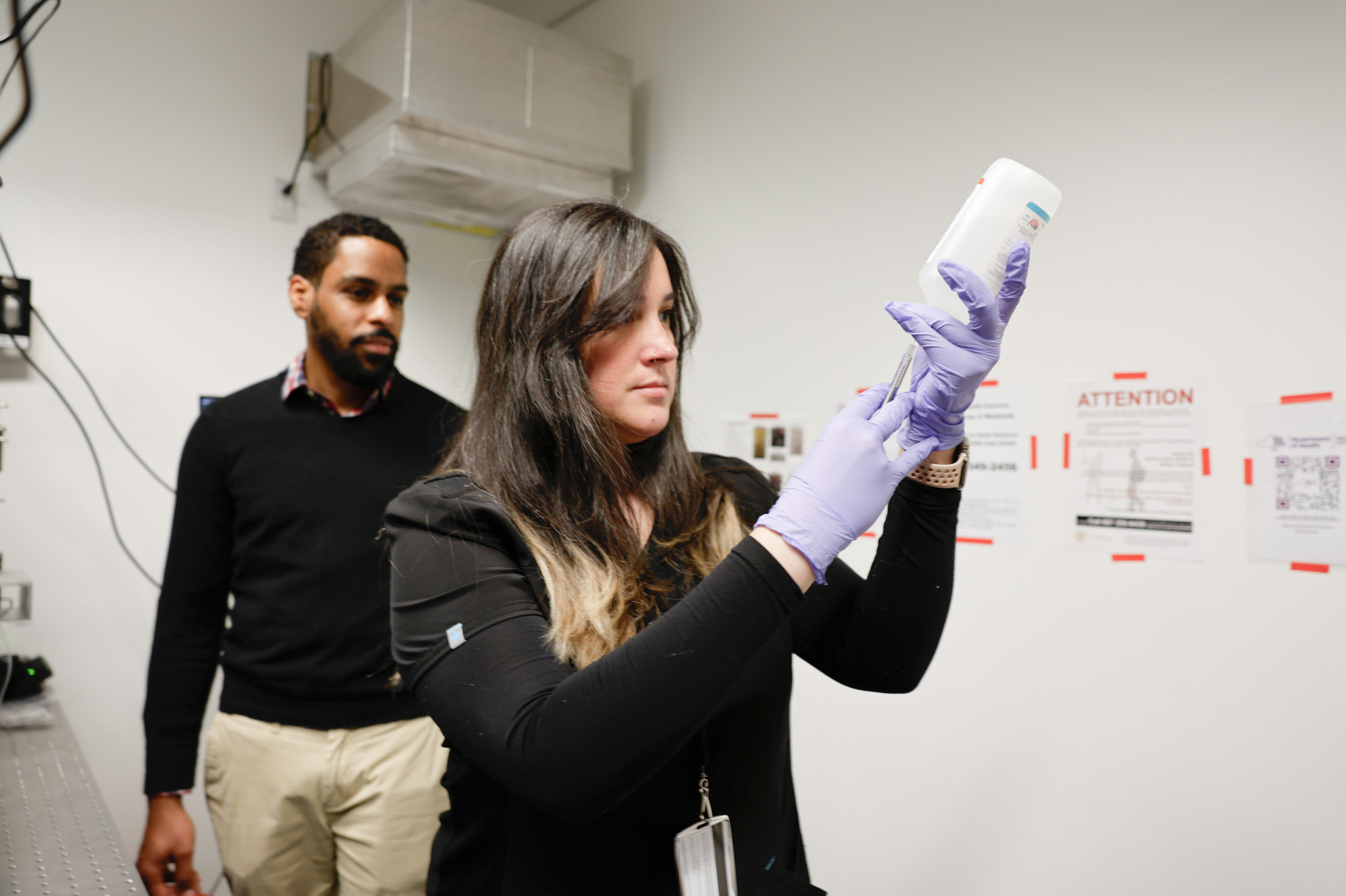 A researcher wearing purple gloves extracts liquid from a plastic bottle using a syringe while professor Karl Lewis watches.