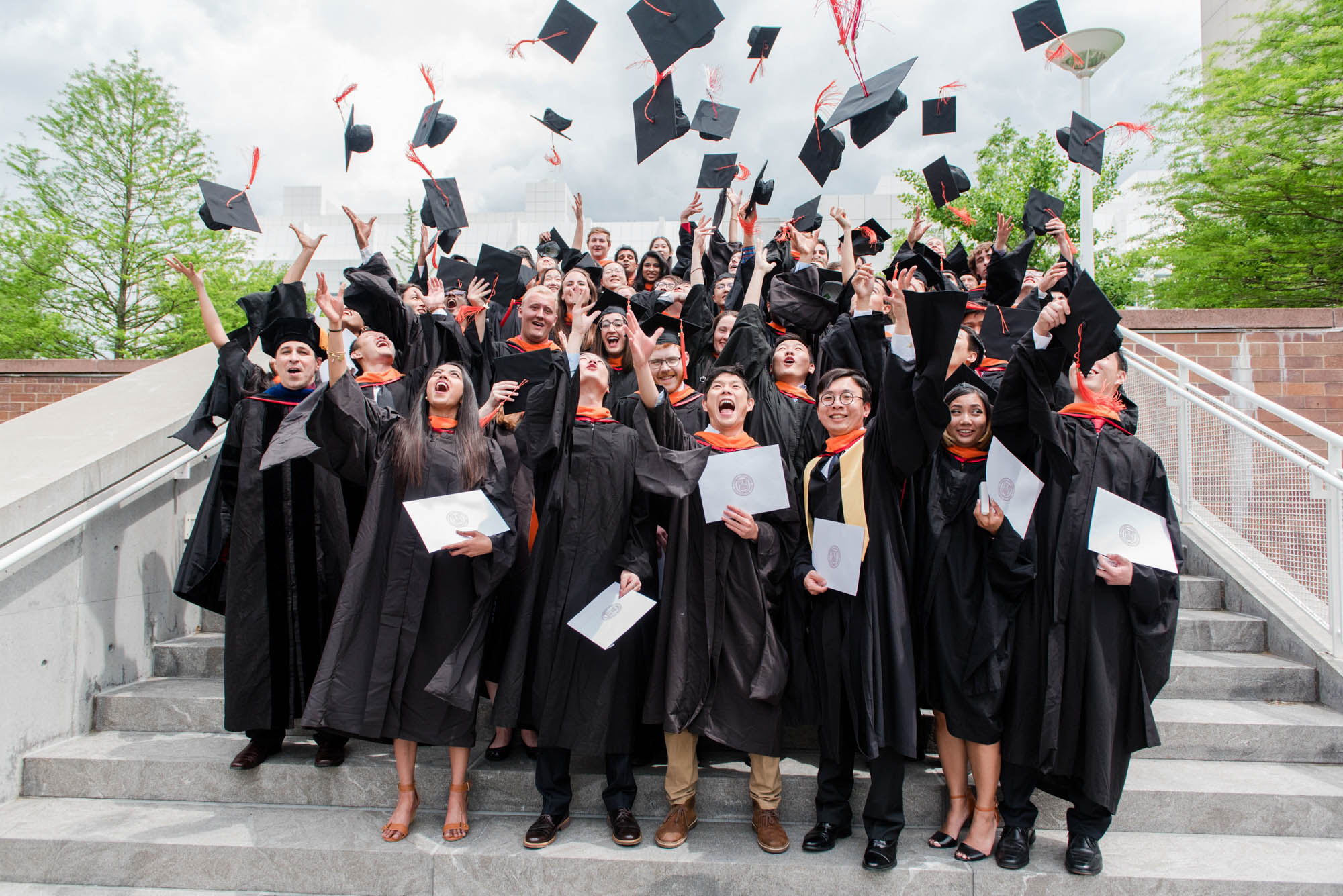 A group of M.Eng. students in graduation regalia toss their caps into the air on the stairs of Weill Hall