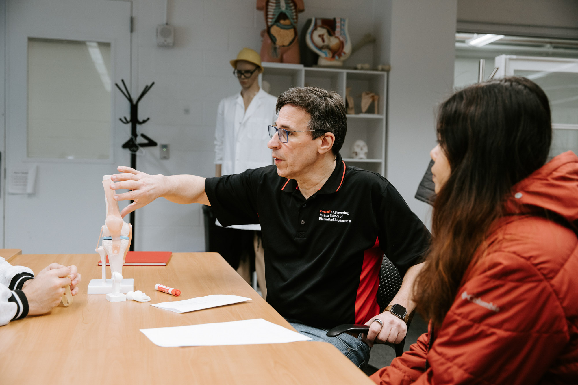 Professor Newton DeFaria points to a model of the human knee as he talks with two students in one of the BME project teams spaces.