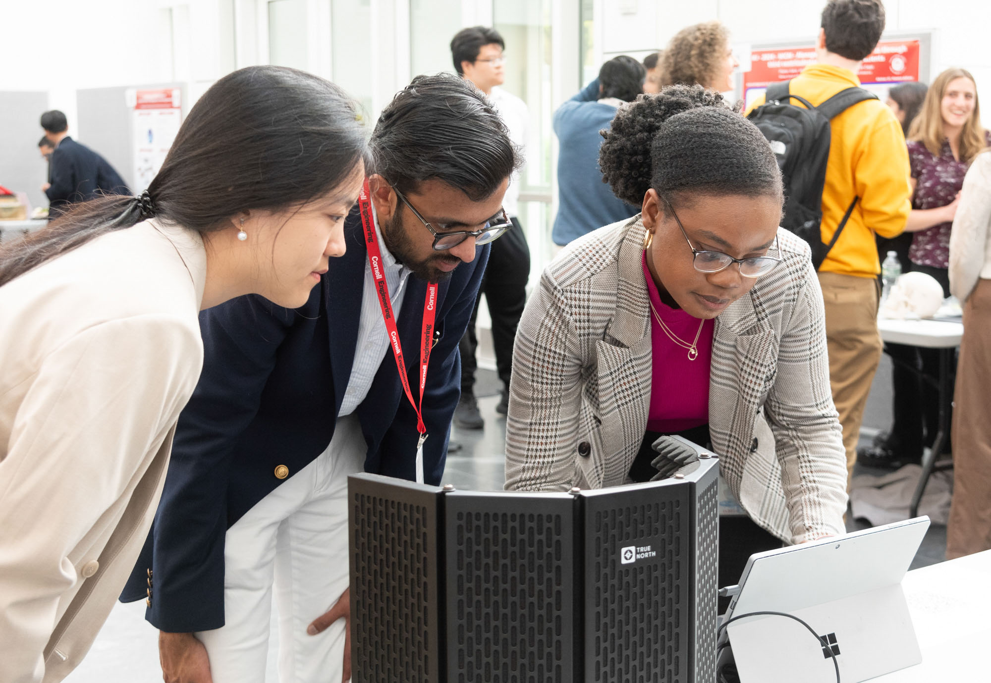 Three students in business attire work on a laptop while other students mingle in the background at the BME Industry Day M.Eng. Showcase.