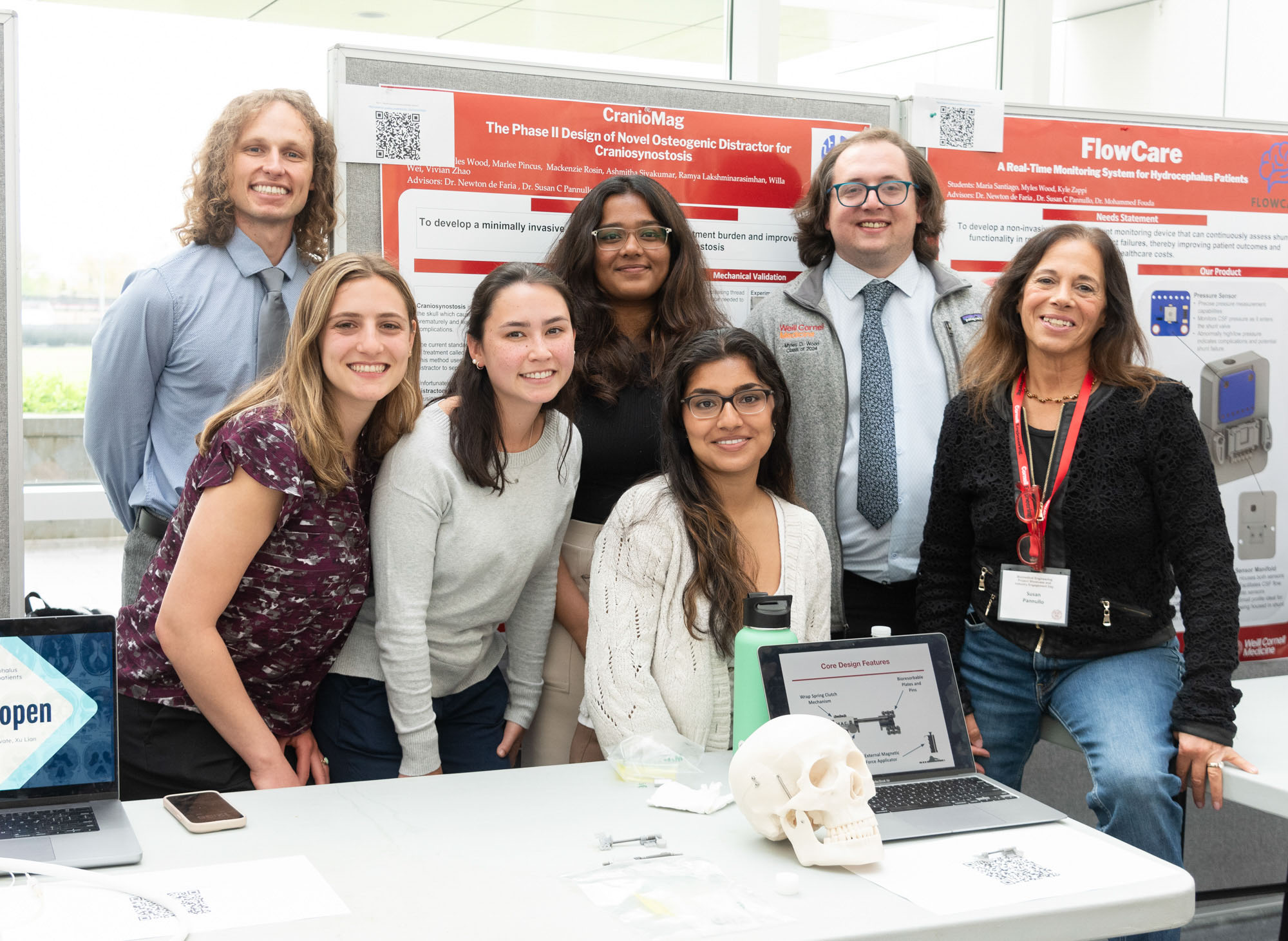 Six students in business attire pose behind a table with a laptop and a model of a human skull on it and in front of their poster with a faculty advisor at the BME Industry Day M.Eng. Showcase.