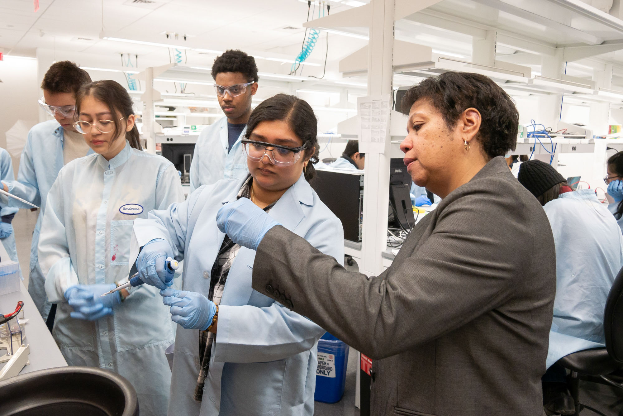 Professor Shivaun Archer wearing blue gloves shows a slide to a group of students wearing lab coats and blue gloves