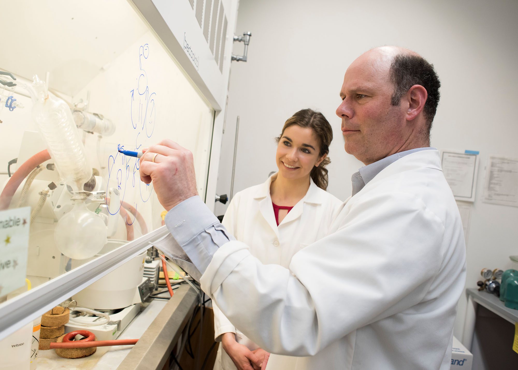 David Putnam with graduate student, wearing lab coats and writing chemical formulas on fume hood glass in lab