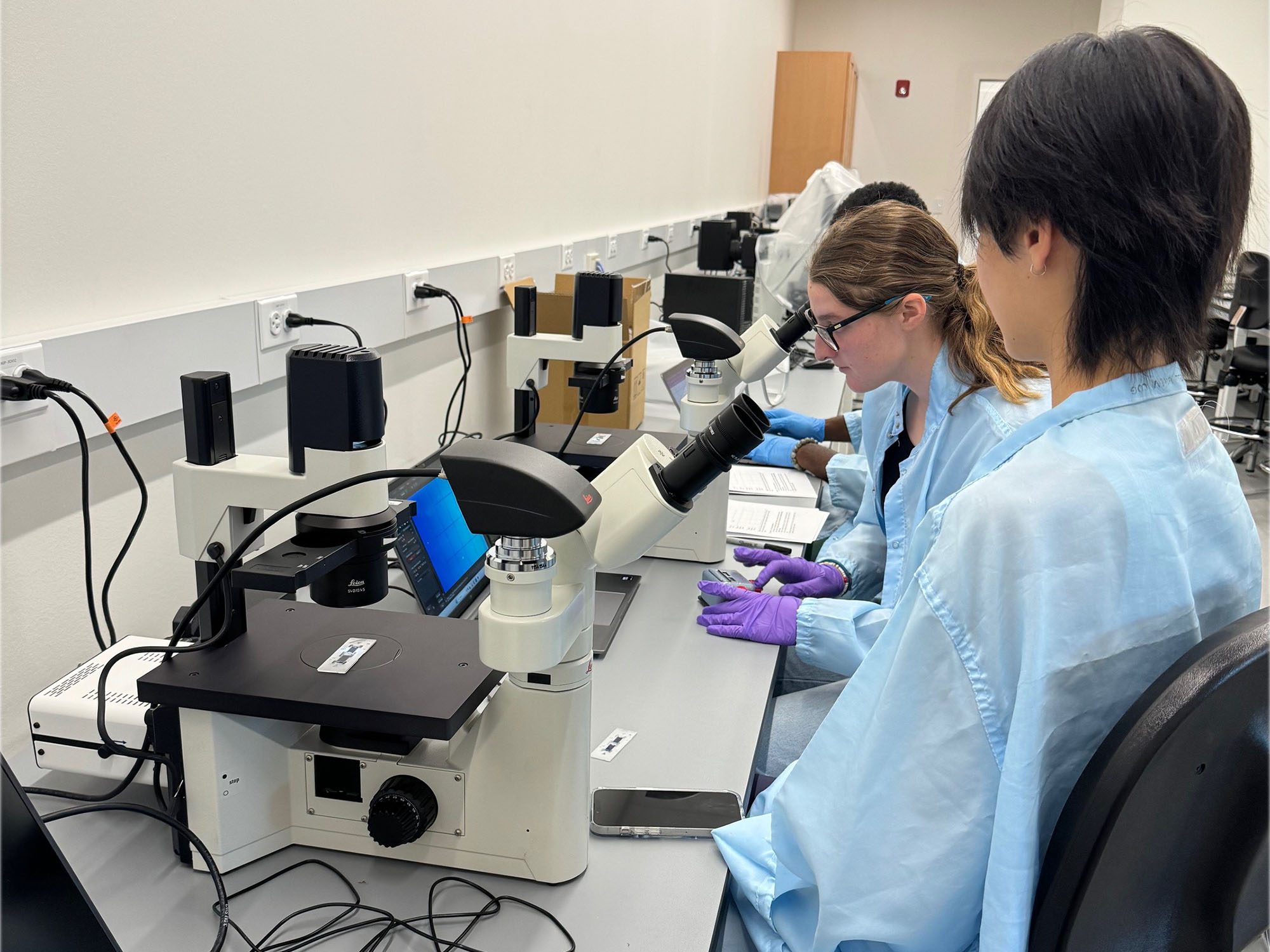 Students wearing lab coats in Tang Hall microscopy lab work at bench with microscopes to analyze cells and tissues.