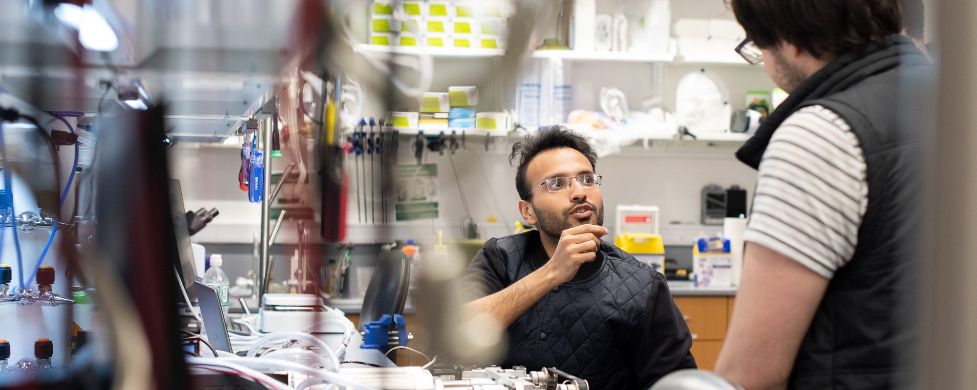 Two students talk with each other in a lab filled with equipment.