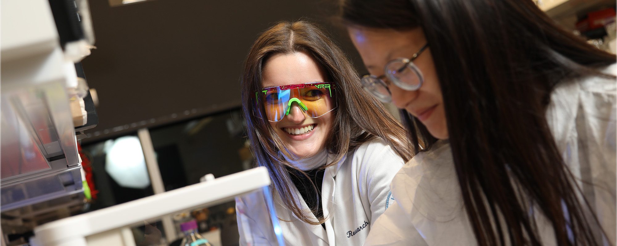 Karl Lewis lab students chat in lab coats and safety glasses.