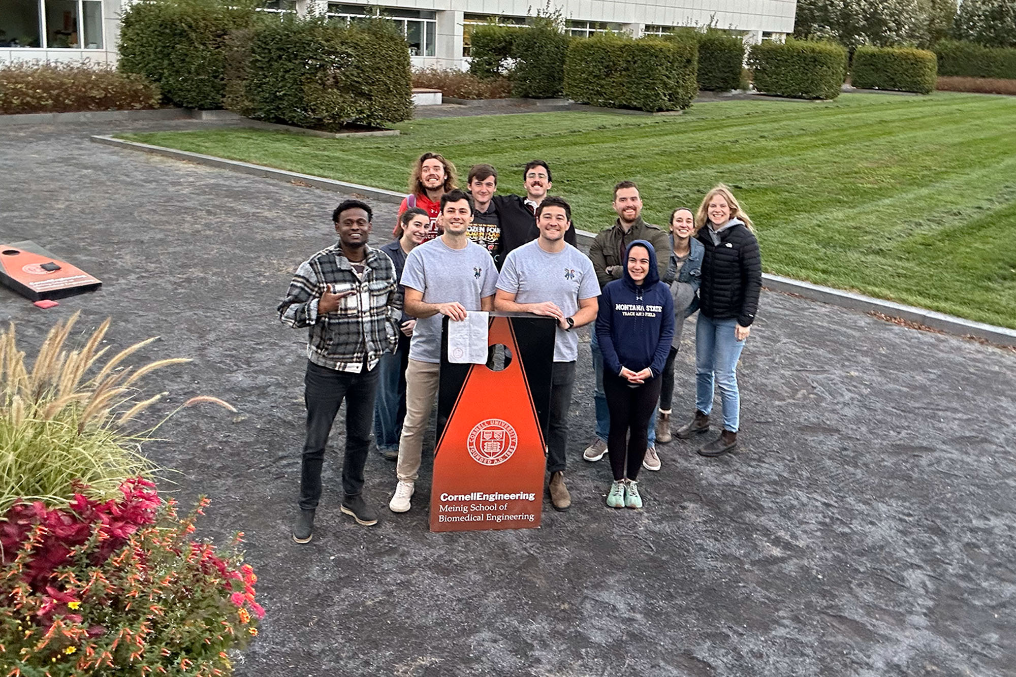Graduate students stand together with corn hole board beside Weill Hall courtyard.