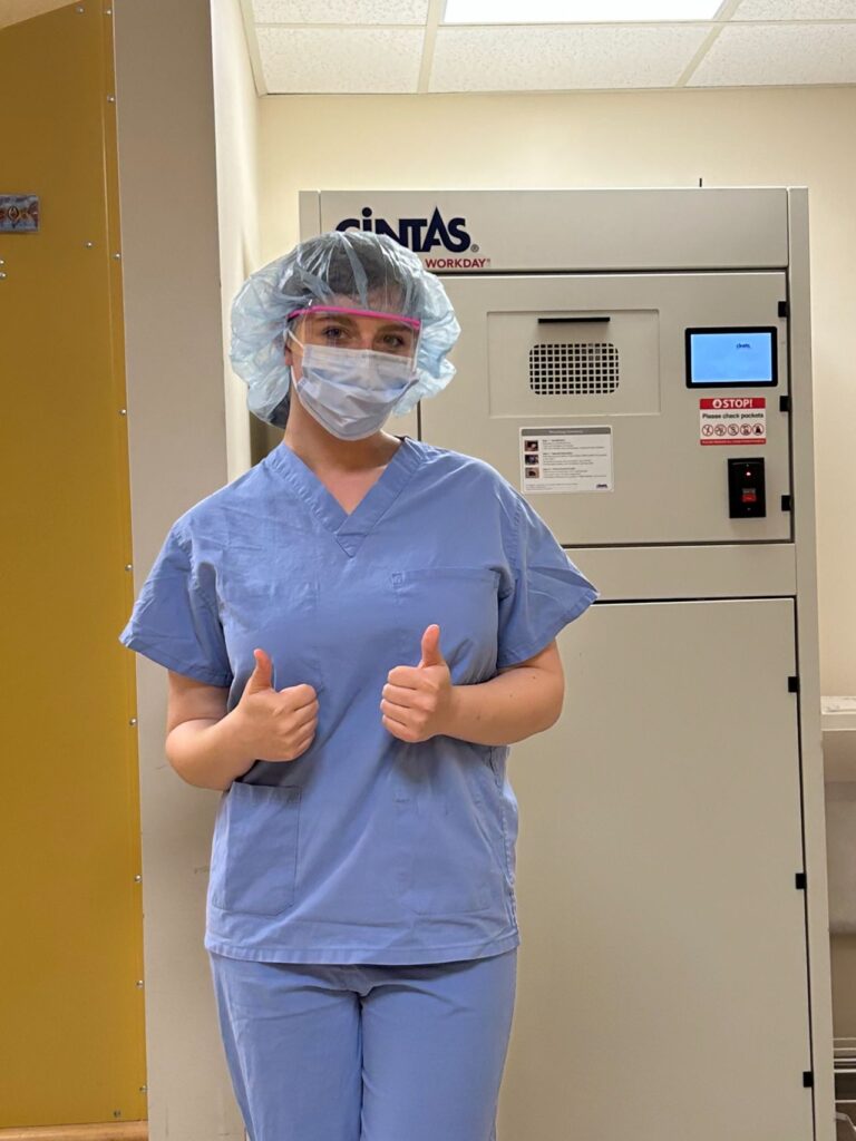 MEng student Maddie Slavett wearing scrubs stands with equipment behind her and thumbs up  at her preceptorship at Guthrie Medicine in Pennsylvania.