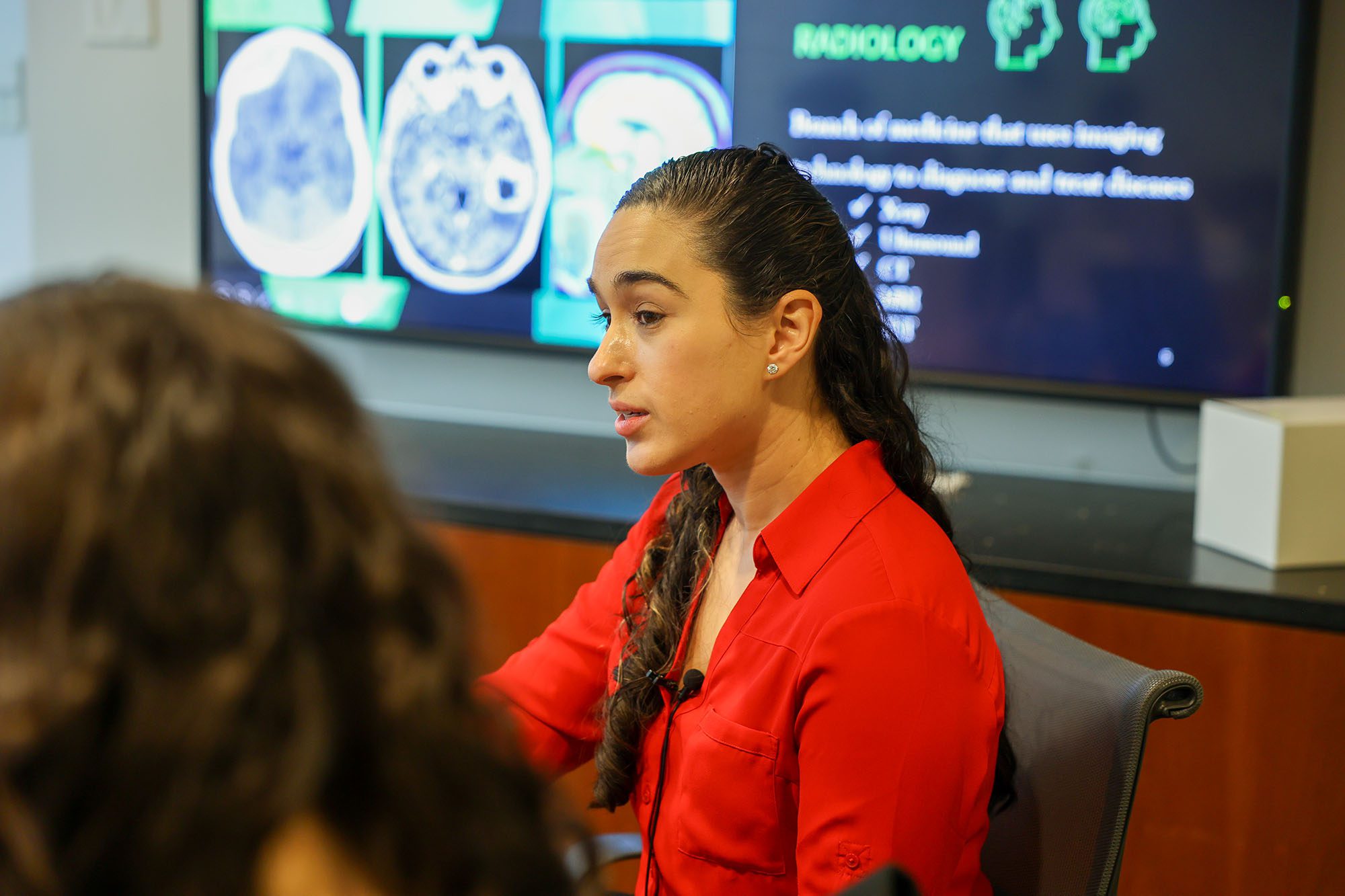 A Weill Cornell physician sits at a table talking to undergraduate students with brain imagery on the screen behind them.