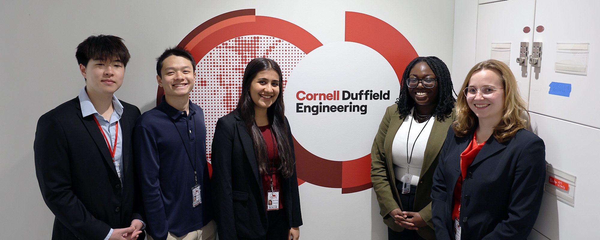 An M.Eng. design team stands together beside a Tang Hall wall decal that says Cornell Duffield Engineering.