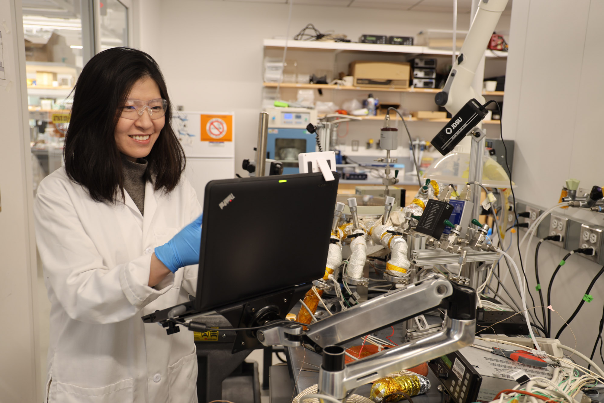 Professor Rong Yang smiling wearing a white lab coat, safety goggles and blue gloves working in her lab