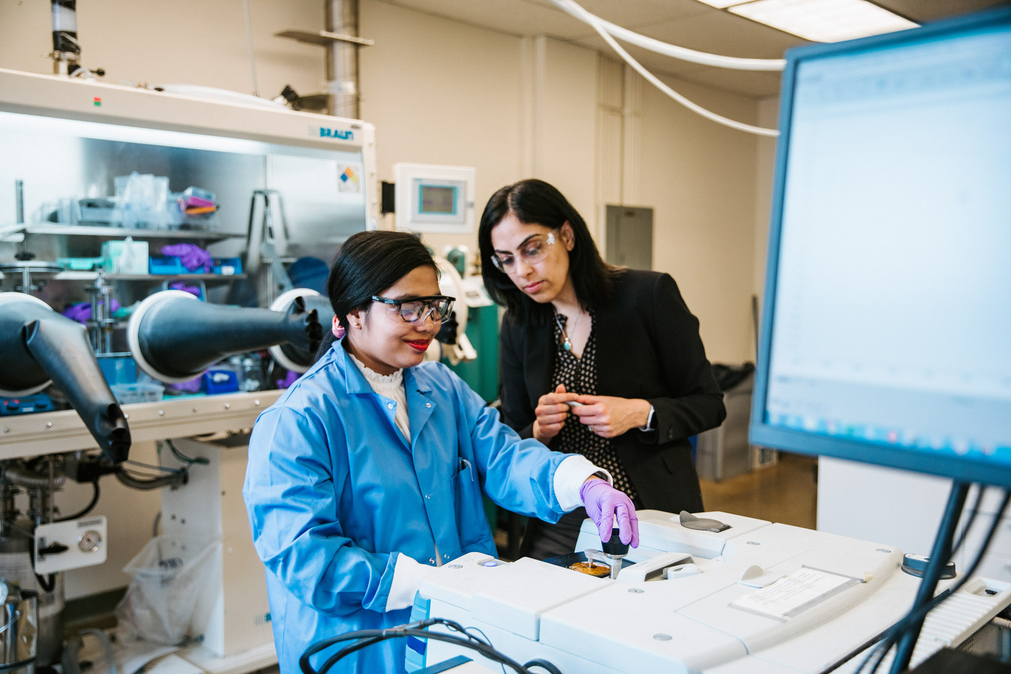 A student wearing a blue lab coat and purple gloves works in the lab while professor Vibha Kalra watches