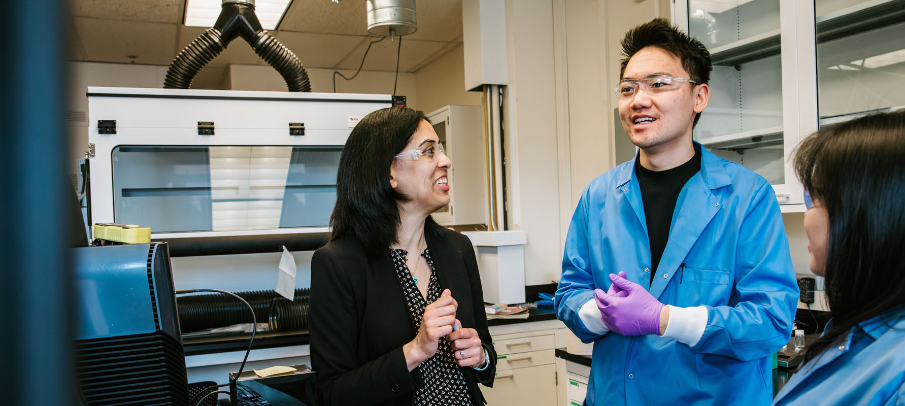 Two students in blue lab coats and safety goggles talk with professor Vibha Kalra in the lab