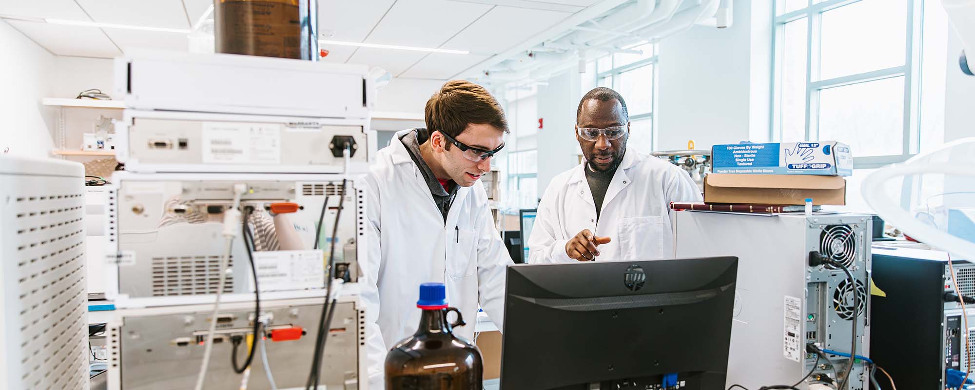 Professor Chris Alabi and a student, both wearing lab coats, looking at a computer monitor in the Alabi Lab