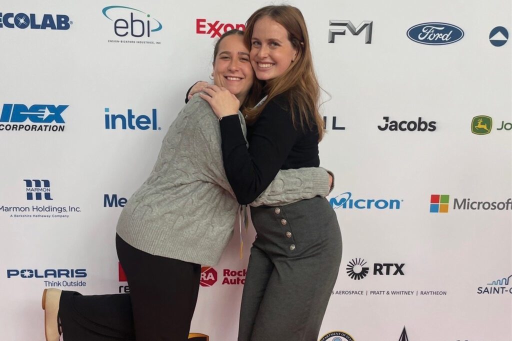 Julia Salatti hugs her co-President in front of a logo backdrop at the Society of Women Engineers (SWE) national conference.
