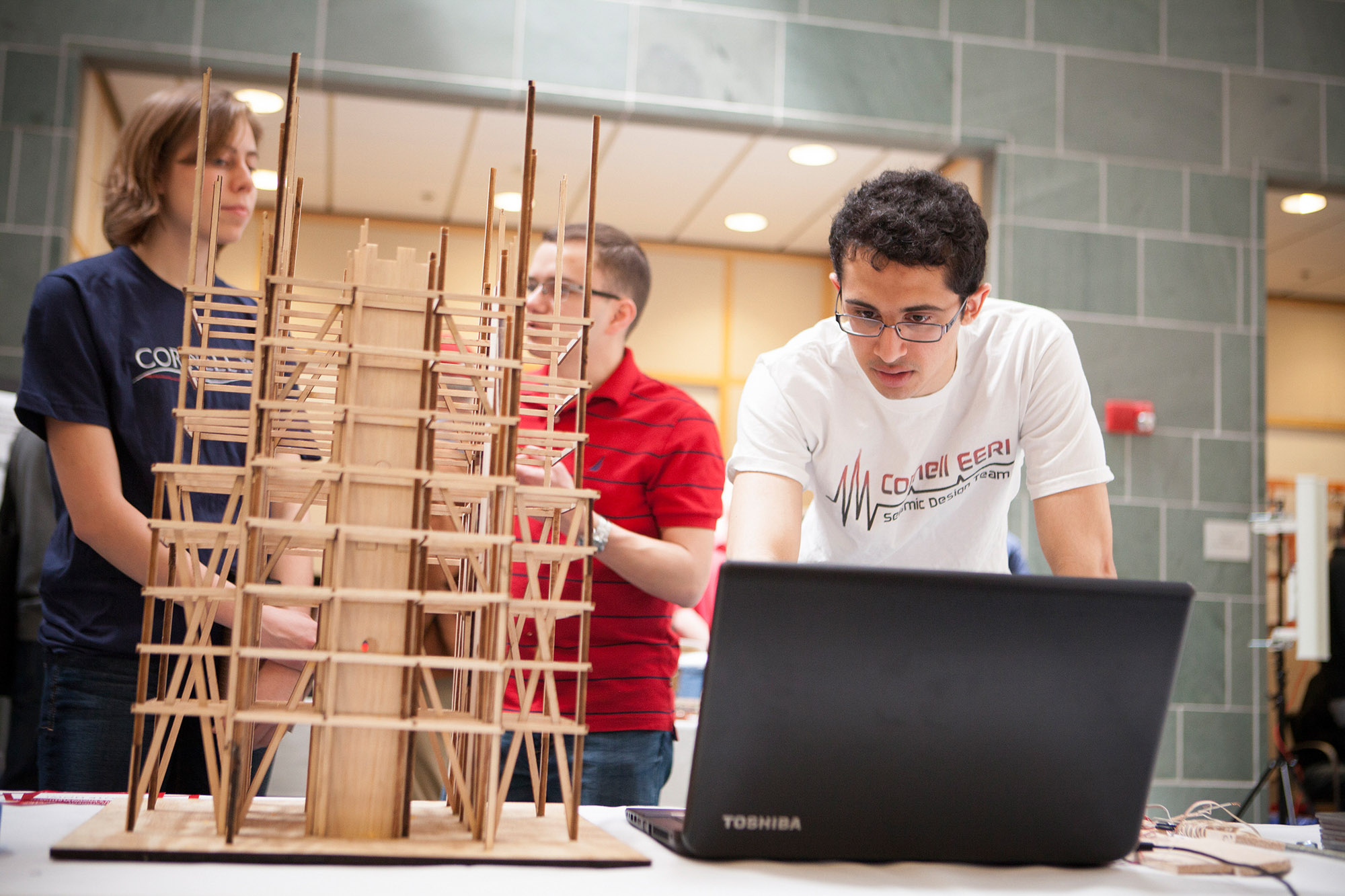 Student looks at computer next to tower made out of wood as two students stand in background.
