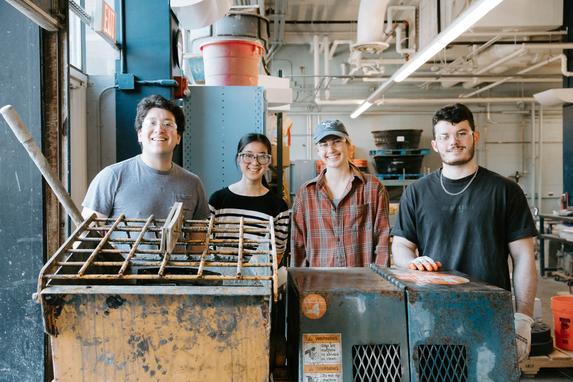 Four smiling students wearing protective glasses stand behind metal equipment