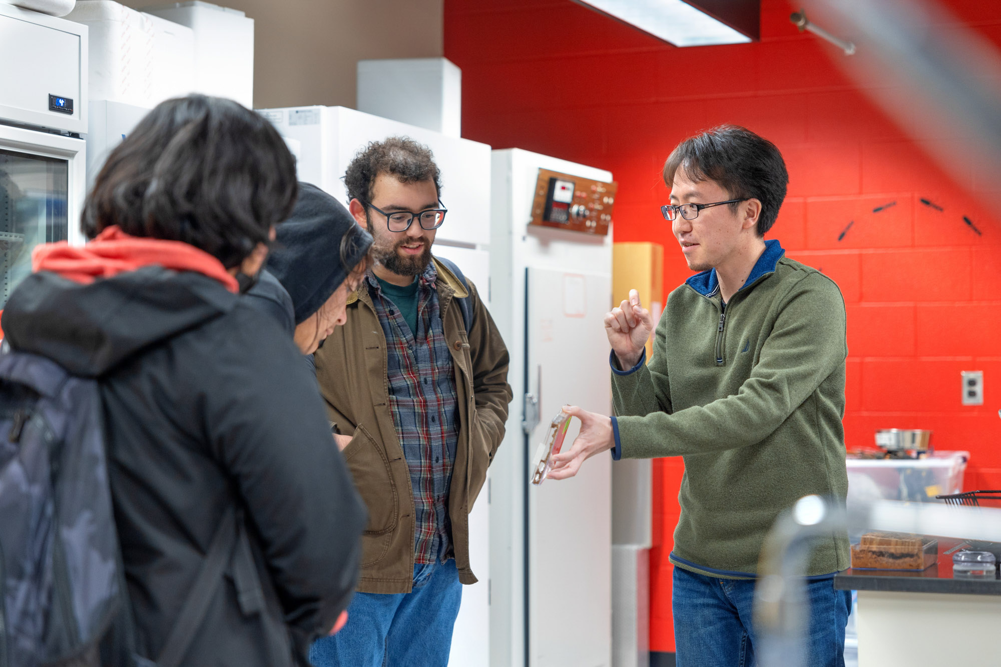Two Cornell researchers talk to two high school students in an entomology lab. One researcher is holding a clear box with termites in it.