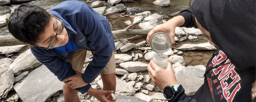 Two students stand in a creek bed and put water into jars.