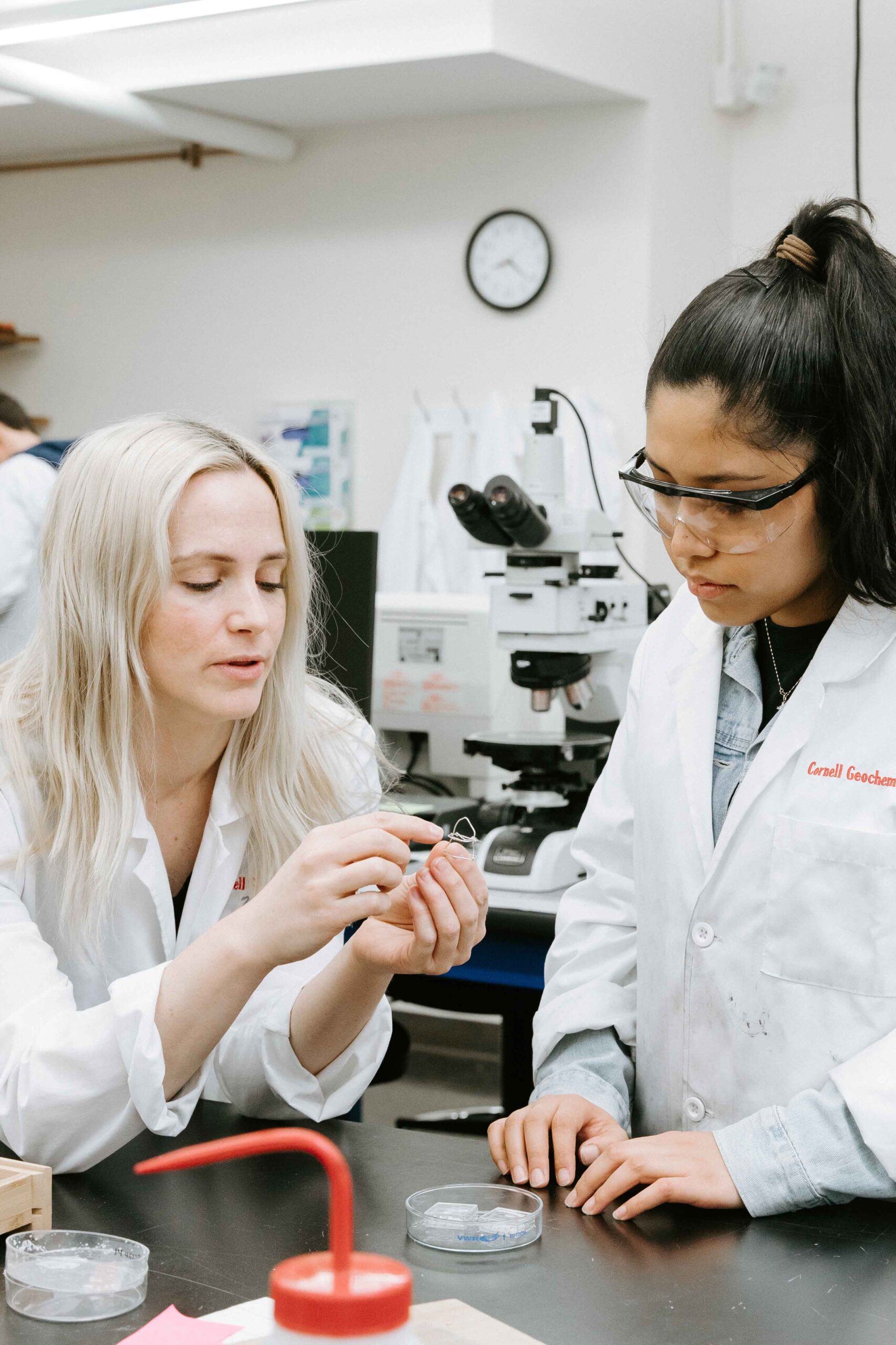 Megan Holycross and a student talk while wearing lab coats and looking at a small piece of equipment.