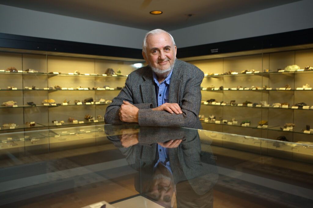 William Bassett leans on glass table, smiling, with glass shelves of minerals and gems in background