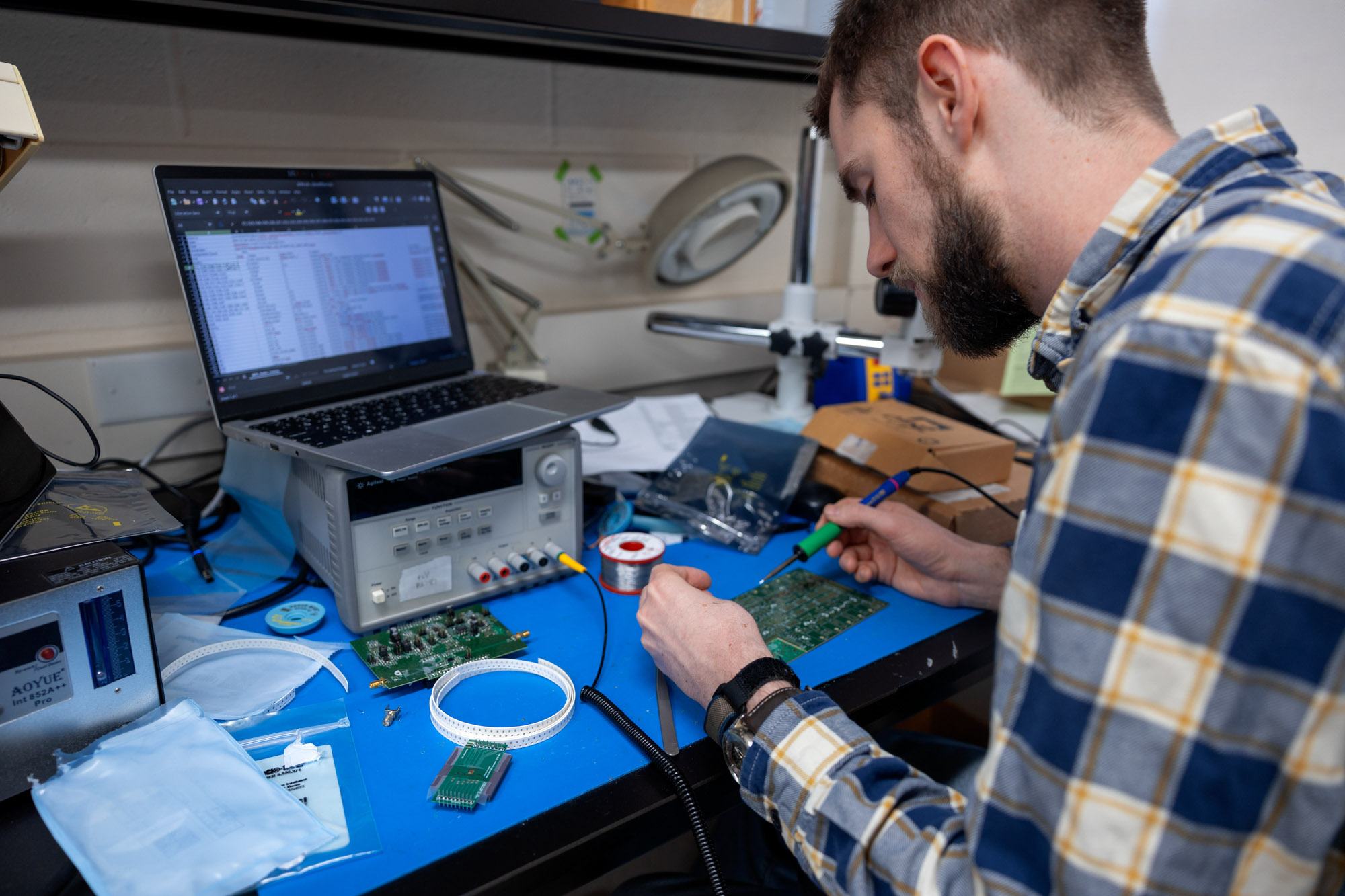 Student works on circuit board while holding a soldering iron in front of laptop, displaying a spreadsheet of data.