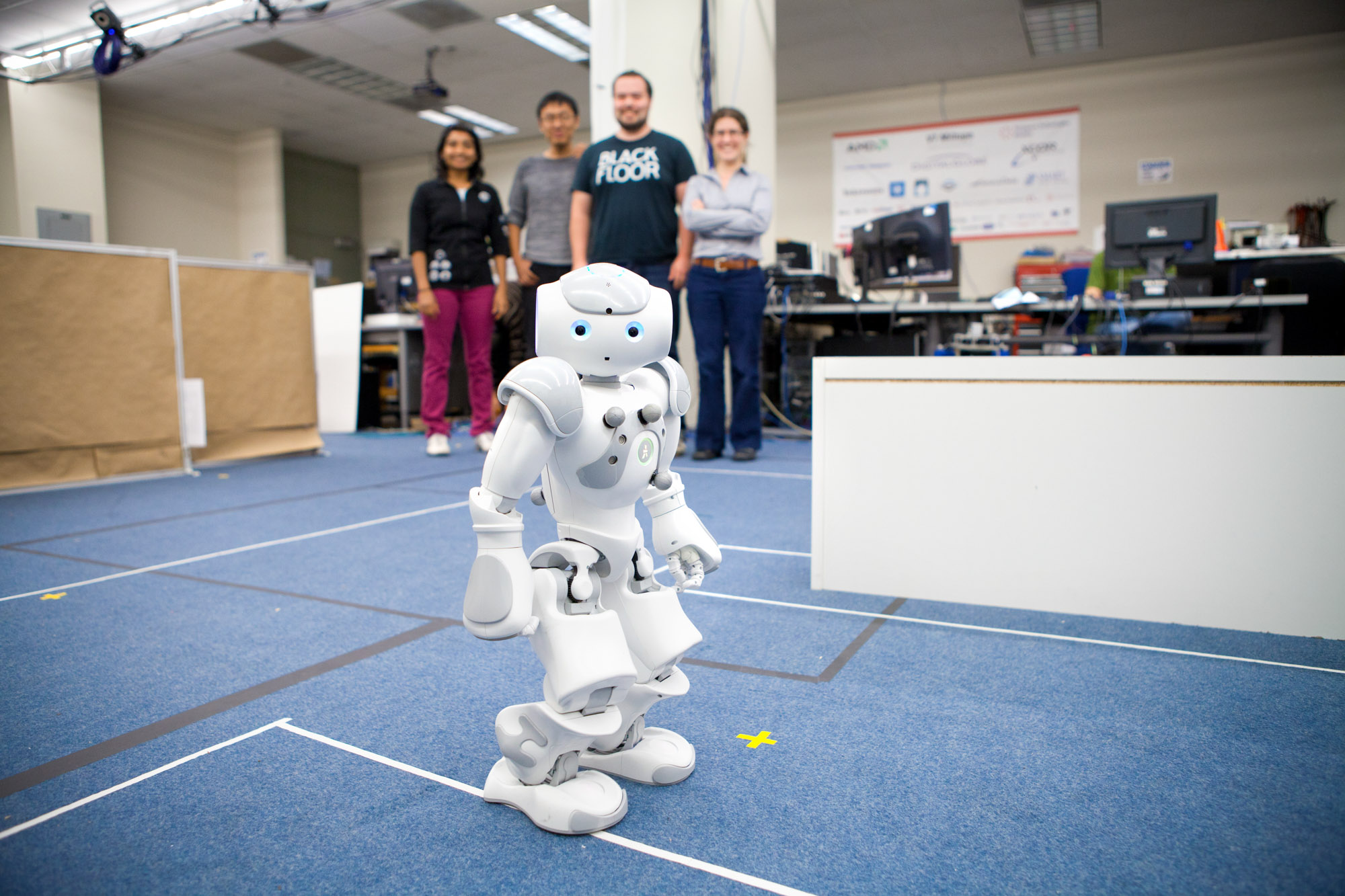 A small white humanoid robot stands on blue carpet in the autonomous systems lab in Upson Hall. Hadas Kress-Gazit and three others are out of focus in the background.