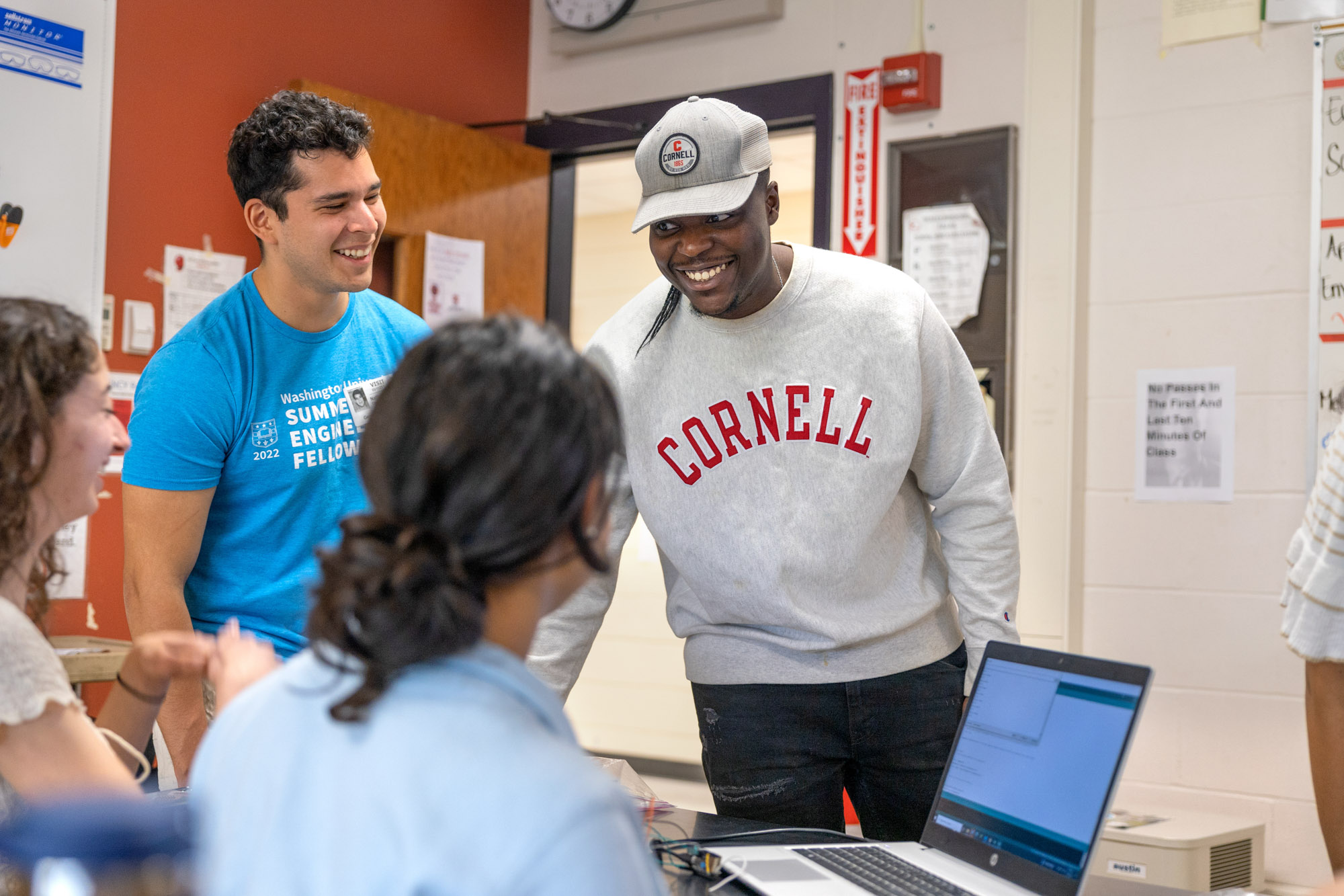 a student wearing a Cornell sweatshirt and hat smiles in front of three other students who are also smiling