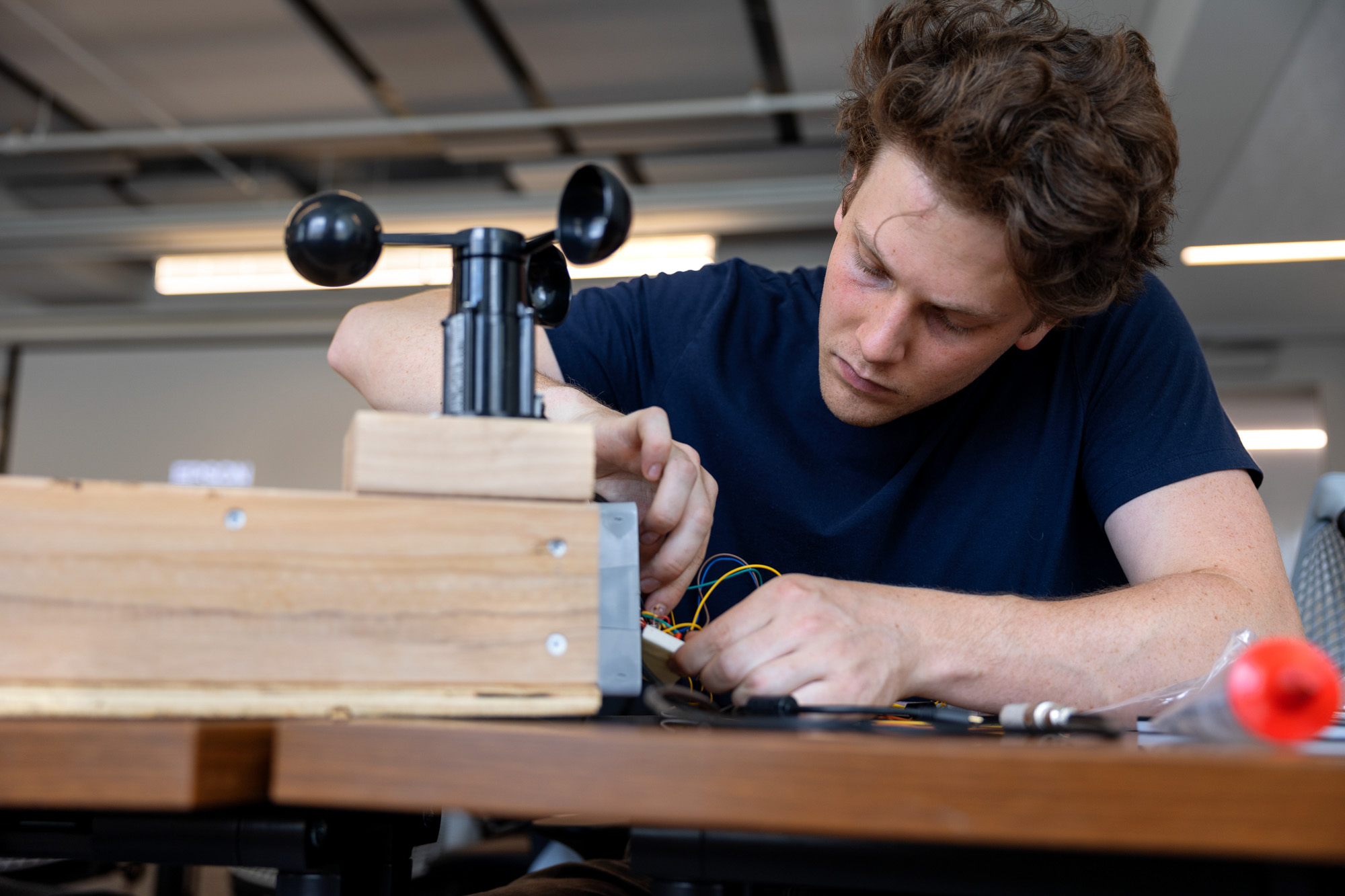 a student in a blue shirt works on a device with yellow and blue wires hooked up to a black wind vane
