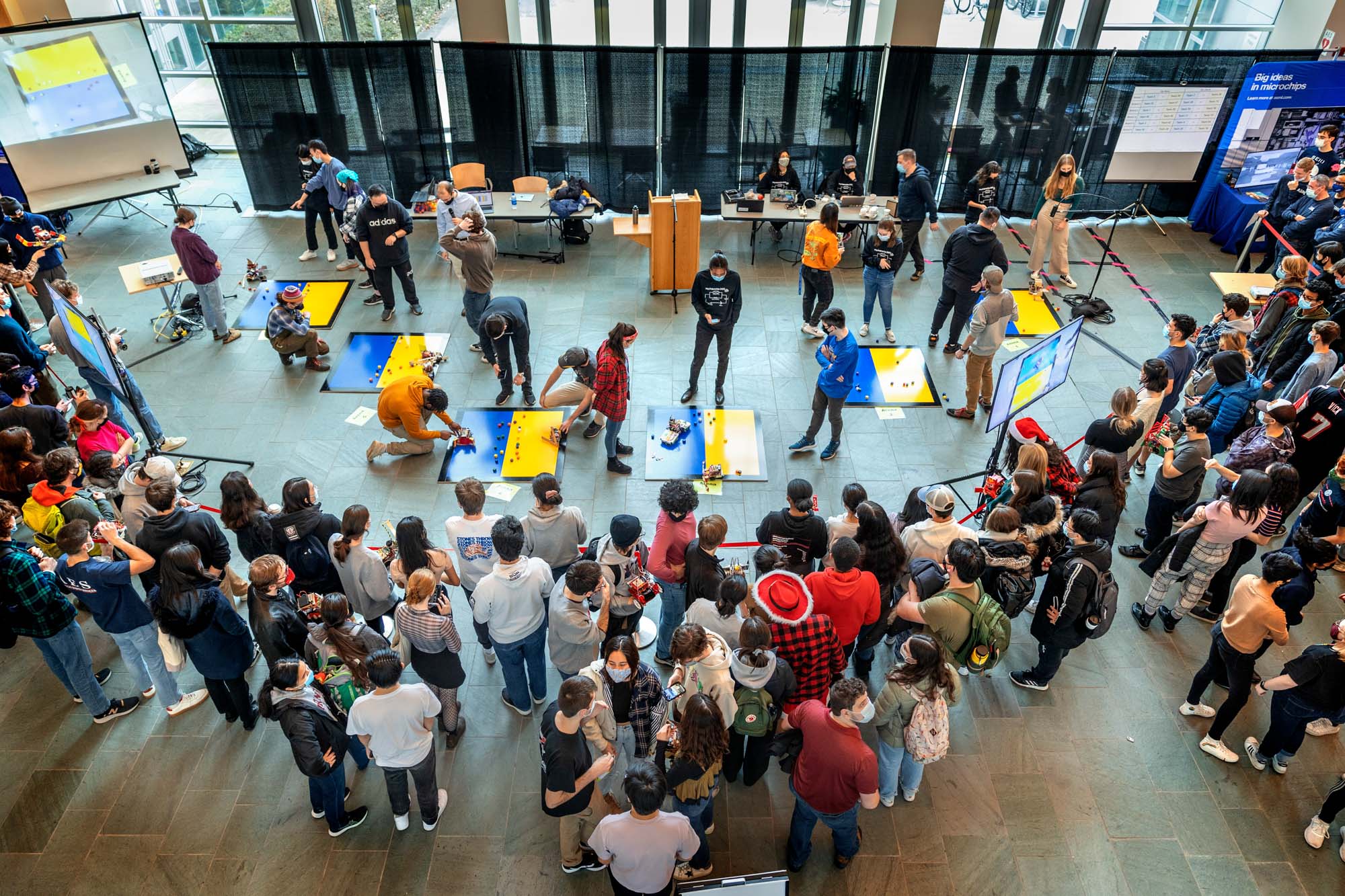 Aerial view of 50 students in Duffield Atrium, gathered around a space with six blue-and-yellow squares on the floor that will be used as playing fields for the robotics competition.