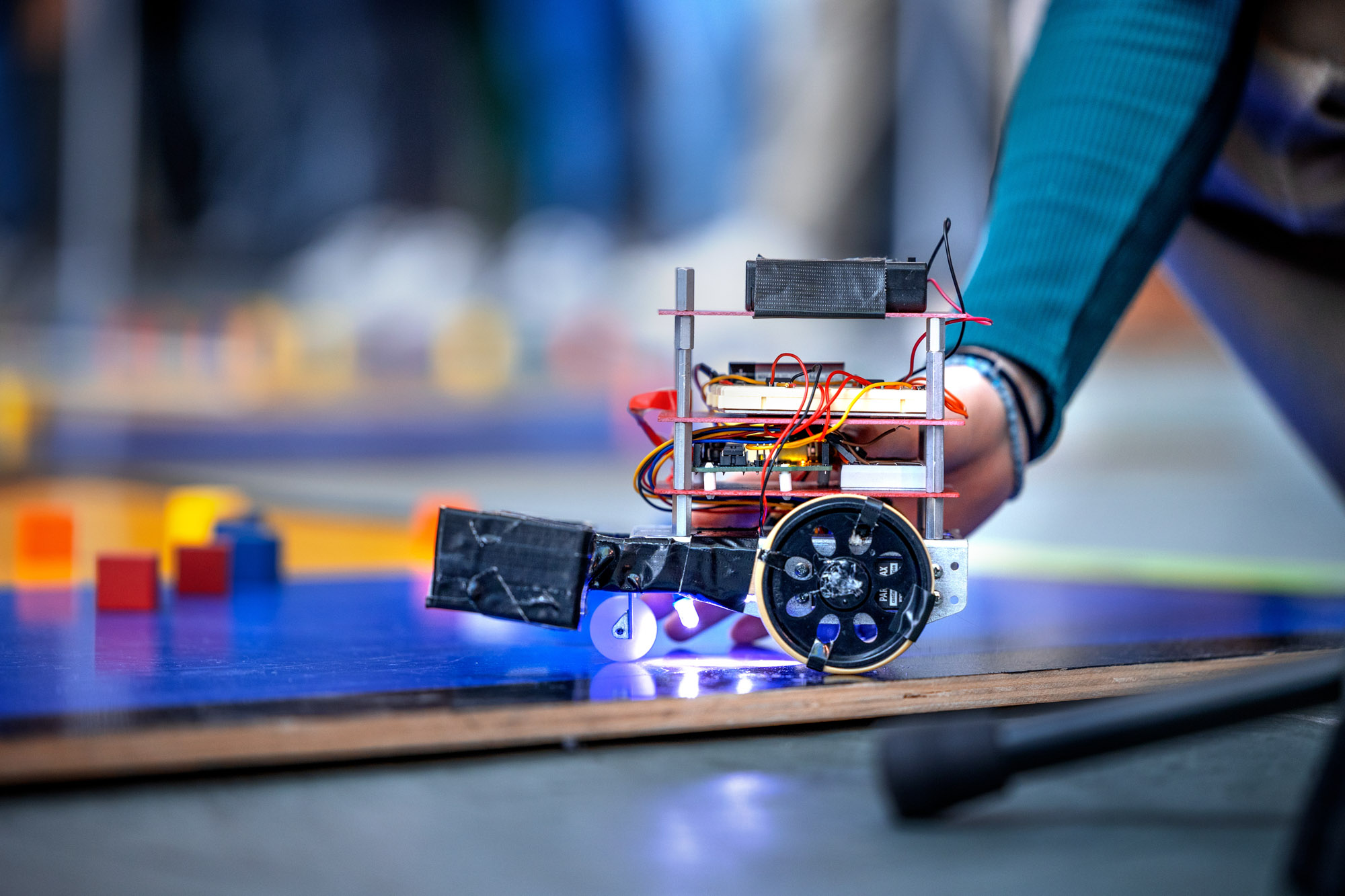 Close-up of a wheeled robot on a blue board. A hand and arm a visible in the background, touching the robot.