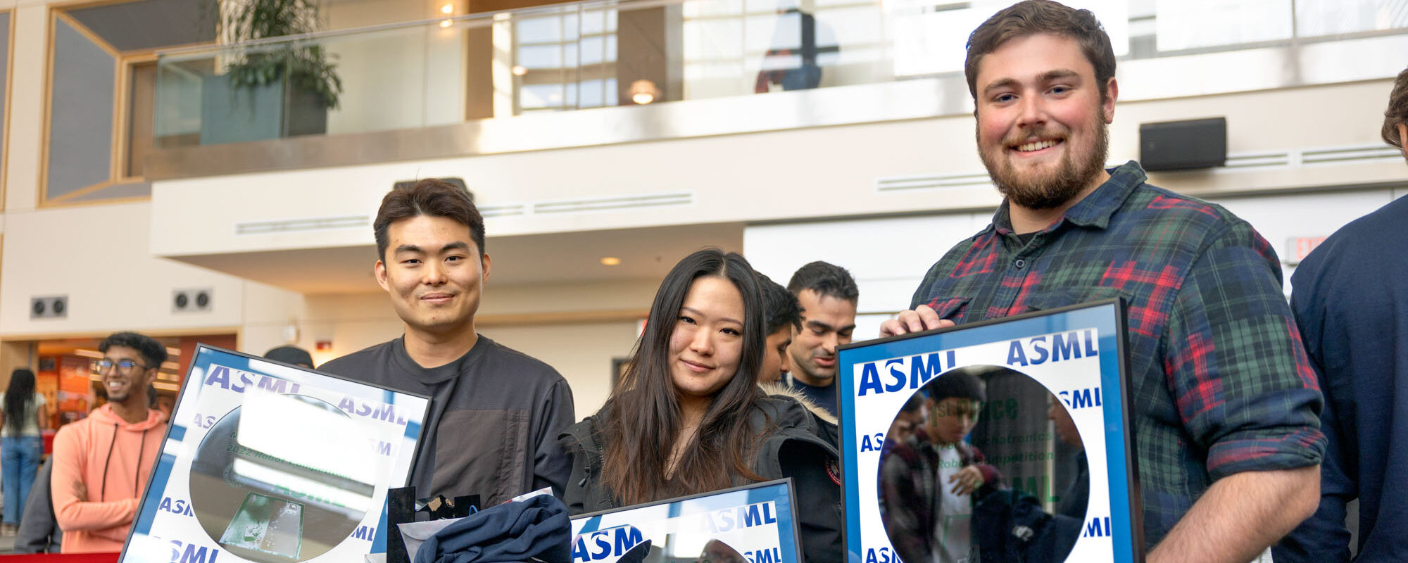 Three students hold framed trophies composed of semiconductor metal and the letters ASML inside the Duffield Hall atrium