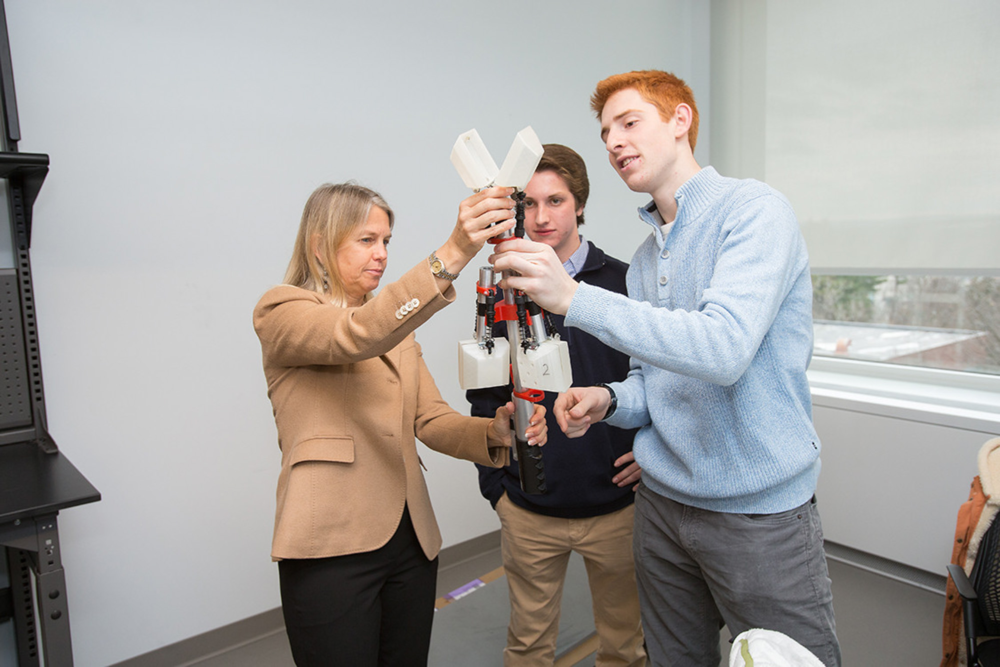 Dava Newman holds and observes a metallic and plastic device with two students inside a classroom.
