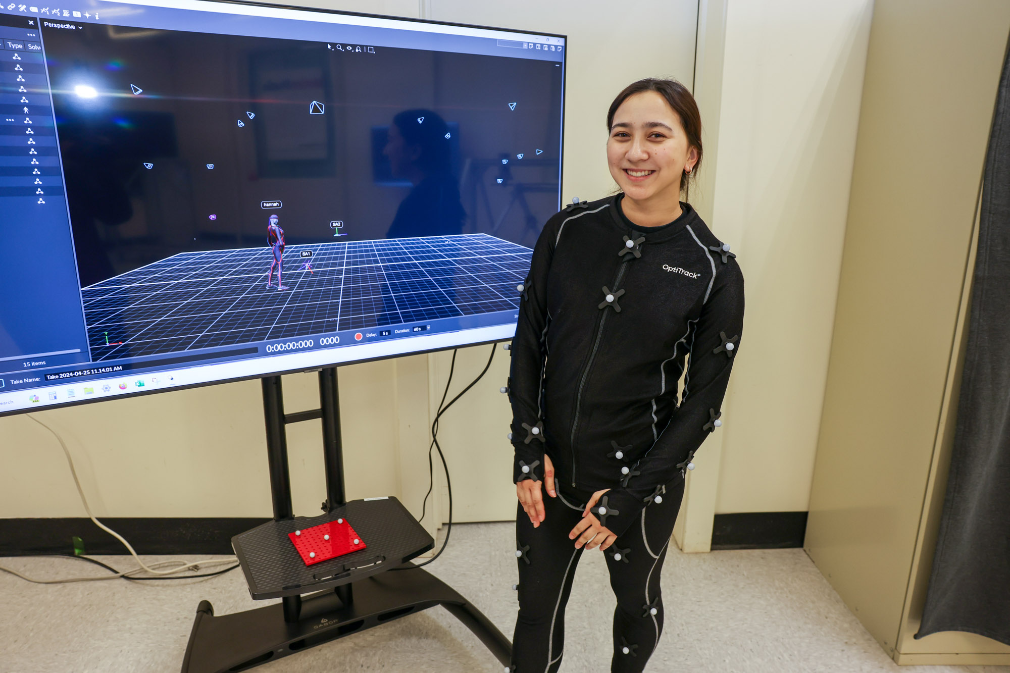 A woman in a black bodysuit smiles with a large television monitor in the background.