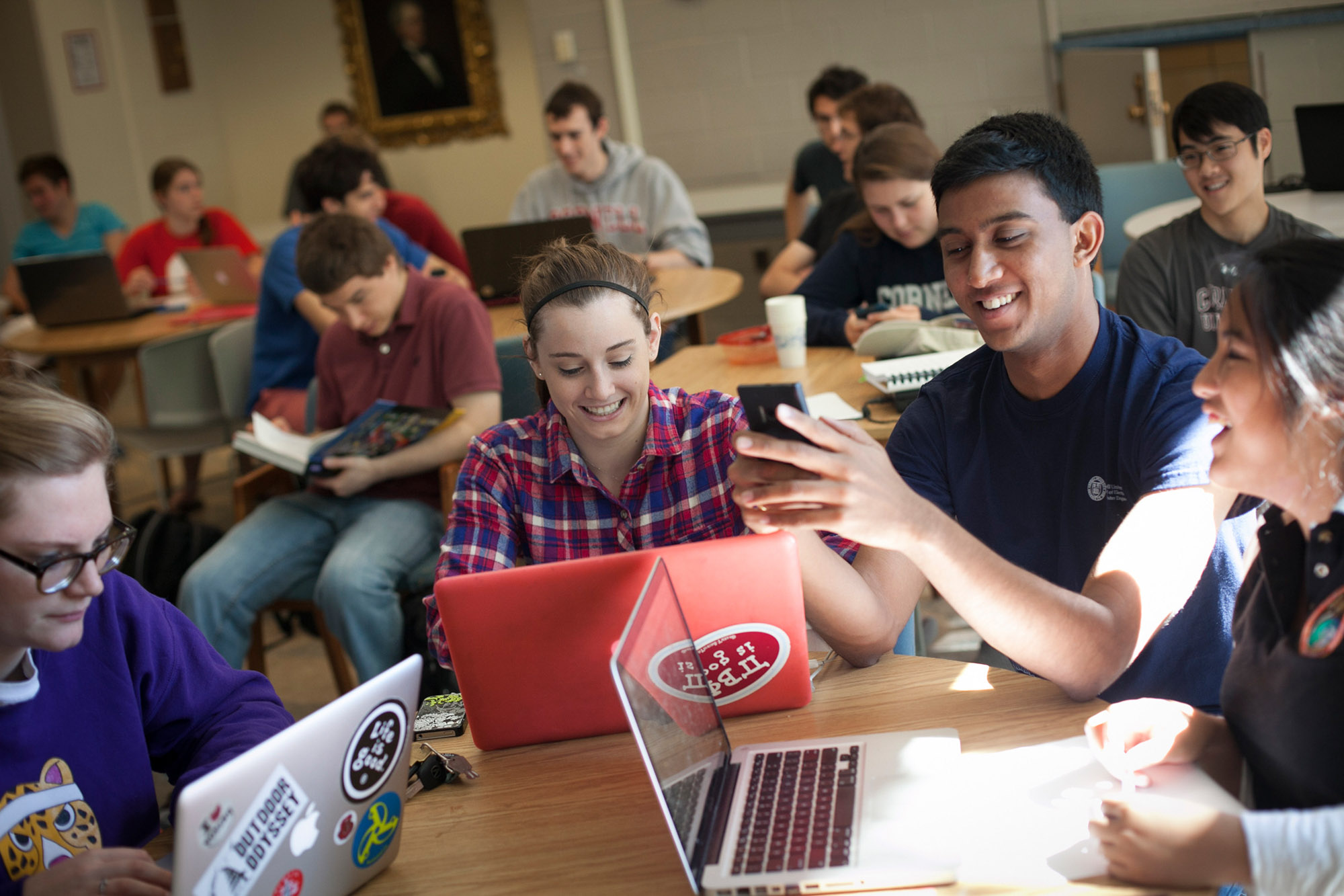 students sit at tables with open books, laptops, and phones.