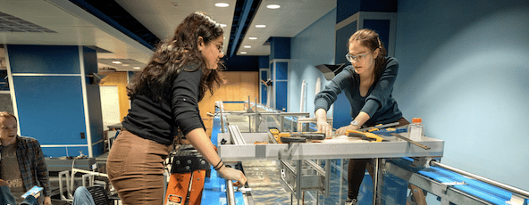 two students stand on ladders to allow them to adjust machinery in a flume test apparatus