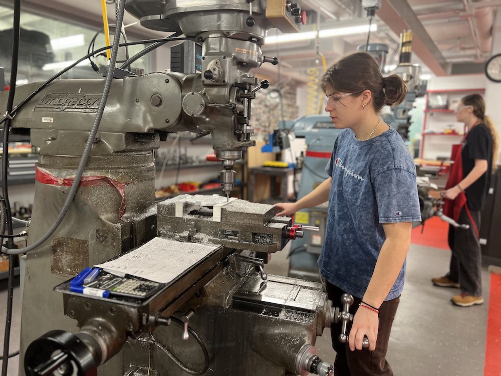 Student wearing safety glasses adjusts knob on a drill press.