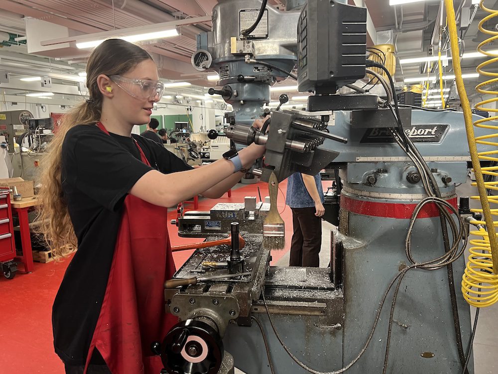 Student wearing safety glasses and ear protection makes adjustments to a drill press.
