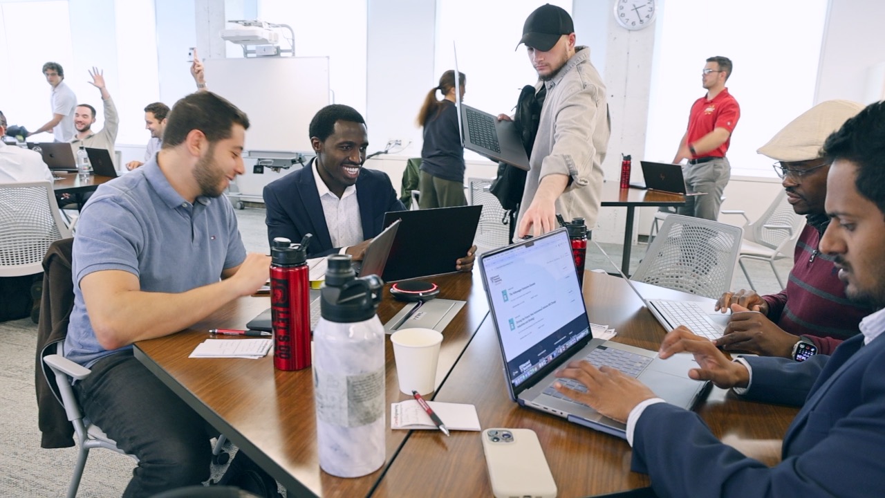 five engineering management students work together at a table using laptops, pens, and paper.